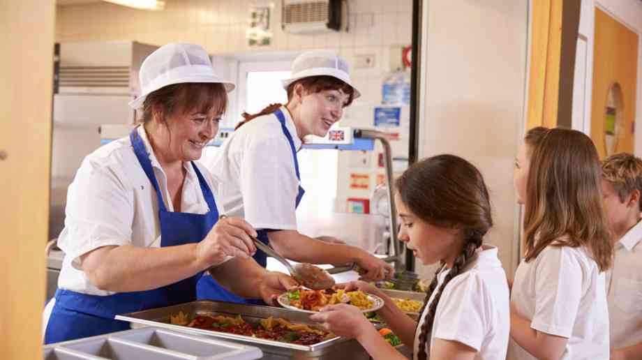 Two dinner ladies serve lunch to students queueing in a school canteen.